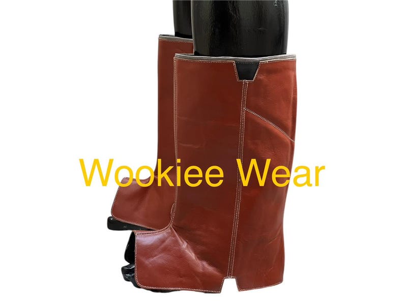 May include: Brown leather chaps with white stitching and the text "Wookiee Wear" in yellow. The chaps are knee-high and appear to be new. The leather has a slight sheen.