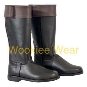Puede incluir: Un par de botas de montar de cuero negro con la parte superior de cuero marrón. Las botas son hasta la rodilla con un diseño clásico. El texto "Wookiee Wear" es visible en amarillo en la imagen.
