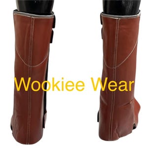 May include: Pair of brown leather boots with white stitching and black buckles. The boots have a silver trim at the top and the text "Wookiee Wear" in yellow.