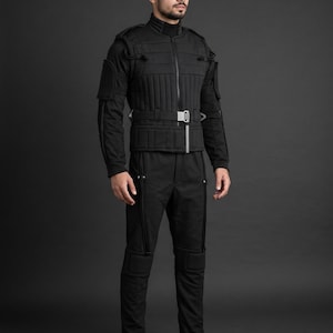May include: A black tactical-style jacket and matching trousers set. The jacket features vertical quilted padding, a high collar, a front zip, and a waist belt with a silver buckle. The trousers include reinforced knee panels and vertical piping.