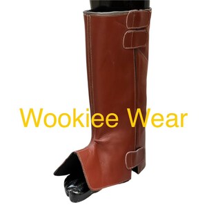 May include: A brown leather boot with two buckle straps and white stitching. The boot features a protective front flap and is designed to cover the lower leg. The text "Wookiee Wear" is displayed in yellow.