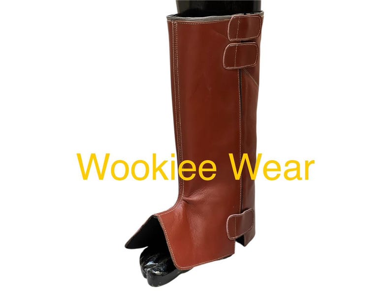 May include: A brown leather boot with two buckle straps and white stitching. The boot features a protective front flap and is designed to cover the lower leg. The text "Wookiee Wear" is displayed in yellow.