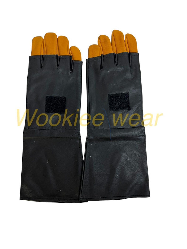 Pu&ograve; includere: Un paio di guanti neri con le punte delle dita arancioni. I guanti hanno un polsino lungo e una toppa in velcro sul dorso della mano. Il testo "Wookiee wear" &egrave; visibile nell'immagine.