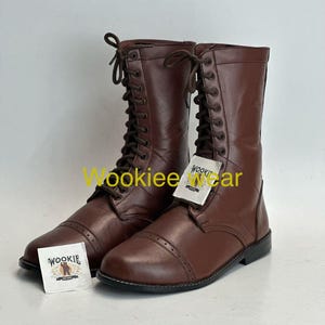 May include: A pair of brown leather lace-up boots with a cap toe design. The boots have a tall shaft and a flat sole. Each boot has a tag that reads "Wookiee Wear".