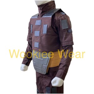 May include: Brown costume with gray accents, featuring a high collar and padded chest. The costume includes a belt and is designed with multiple square patches. The sleeves are long and the gloves are white.