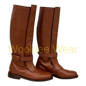 Puede incluir: Un par de botas altas de cuero marrón. Las botas tienen un detalle de hebilla alrededor del tobillo y una suela plana. Las botas están hechas de cuero liso y están diseñadas para usar.