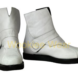 May include: A pair of white leather boots with a black sole. The boots have a unique design with a fold-over flap and a strap with a circular button. The boots are approximately 8 inches tall.