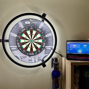 Può includere: Un bersaglio elettronico a tema Star Wars con una cornice nera e un centro bersaglio colorato. Il bersaglio è montato su un muro e un laptop che mostra un punteggio di gioco è nelle vicinanze.