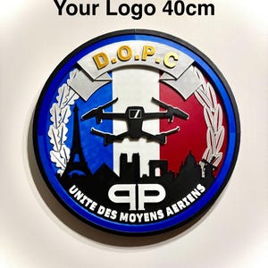 Peut inclure: Une plaque murale circulaire de 40cm avec un fond aux couleurs du drapeau français, une silhouette de drone noire, le texte D.O.P.C et UNITE DES MOYENS AERIENS. Le design inclut les silhouettes de la tour Eiffel et d'autres monuments avec un logo.