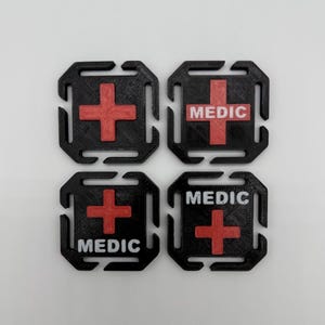 Peut inclure: Quatre patchs carrés noirs avec un motif de croix rouge. Deux patchs portent l'inscription "MEDIC" en blanc. Les patchs ont une forme unique avec des découpes sur les côtés.