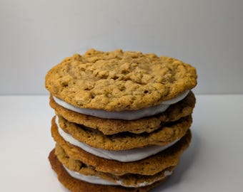 May include: A stack of five oatmeal cream pies. The cookies are golden brown with a textured surface. The white cream filling is visible between each cookie layer. The dessert is stacked on a white surface.