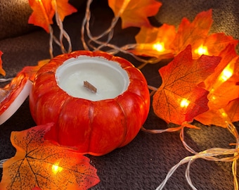 Könnte beinhalten: Eine runde, orangefarbene Kerze in Kürbisform mit weißem Wachs und einem Holzdocht. Die Kerze ist von einer Lichterkette aus orangefarbenen Ahornblättern umgeben, die eine warme, herbstliche Atmosphäre erzeugt.
