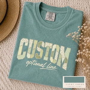 Può includere: Una t-shirt a maniche corte color verde salvia con scollo rotondo ed etichetta del marchio Comfort Colors. La parte anteriore presenta la parola CUSTOM in grandi lettere stampatello con un motivo floreale patchwork, seguita dal testo optional line in corsivo bianco.