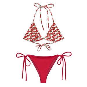 May include: A two-piece bikini set featuring a white halter-style triangle top with a red cherry print and thin tie straps. The matching bottom is a solid red color with thin side-tie closures on both hips, creating a simple and functional design.