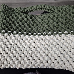 May include: A handmade macrame tote bag in olive green and off-white. The bag features a woven design with a handle at the top. The bag is approximately 14 inches wide and 12 inches tall.