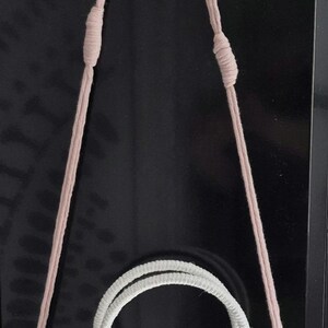 May include: A handmade macrame bag with a white and pink color scheme. The bag features a woven design with a rectangular shape and a circular handle. The shoulder straps are a light pink color. The bag is a stylish accessory.
