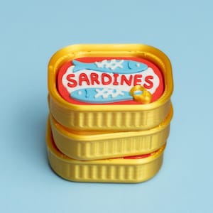 Może przedstawiać: Stos trzech złotych puszek po sardynkach. Na górnej puszce znajduje się czerwona etykieta z napisem "SARDINES" i dwiema niebieskimi ilustracjami ryb. Tło jest jasnoniebieskie.