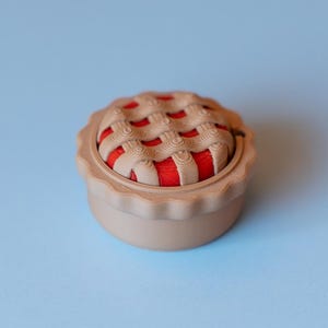 Puede incluir: Un cojín para alfileres beige con forma de tarta y diseño de corteza enrejada. El interior es rojo y el cojín para alfileres está sobre una superficie azul claro. Accesorio de costura decorativo.