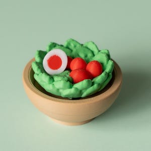 Könnte beinhalten: Eine kleine Holzschale enthält eine leuchtend grüne, salatartige Struktur mit drei kleinen roten Kugeln und einem halbierten Ei mit roter Mitte. Die Schale steht auf einer hellgrünen Oberfläche.