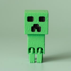 Peut inclure: Une figurine verte et carrée avec une tête carrée, des yeux noirs et une bouche noire. La figurine a une surface texturée et repose sur deux petits pieds. Le fond est de couleur vert clair uni.