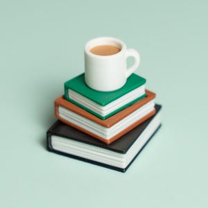 May include: A stack of three miniature books in black, brown, and green, topped with a small white mug filled with a light brown liquid. The books are stacked on top of each other against a light green background.