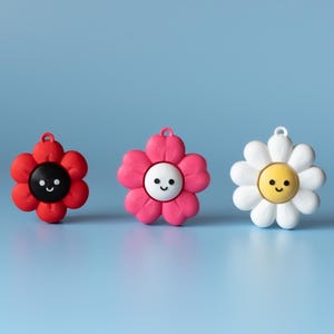 May include: Three flower-shaped charms with smiling faces. One is red with a black center, another is pink with a white center, and the last is white with a yellow center. Each charm has a small loop at the top for hanging. The background is a light blue.