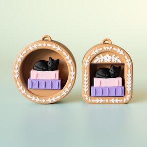 May include: Two decorative ornaments featuring a black cat resting on a stack of books. One is round, the other is arched. The ornaments are tan with white floral accents and the books are pink and purple.