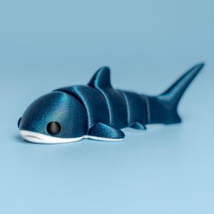 May include: A dark blue shark figurine with a white underbelly and black eyes. The toy shark has a segmented body and fins, and is set against a light blue background.