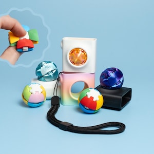 Pode incluir: Uma coleção de brinquedos fidget esféricos em várias cores, incluindo azul metálico, roxo e segmentos multicoloridos. O conjunto inclui um estojo retangular branco e degradê com um recorte circular e uma alça preta.