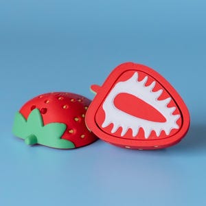 Op de afbeelding: Twee rode aardbeivormige objecten op een blauwe achtergrond. Eén is heel, de andere is doormidden gesneden en onthult een wit interieur met een rood midden. De hele aardbei heeft groene bladeren en kleine gele zaden.