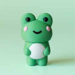 Op de afbeelding: Een groene kikkerfiguur met een witte buik, roze wangen en zwarte ogen. De kikker heeft een lachend gezicht en staat op een lichtgroen oppervlak. Het beeldje is een schattig, grillig ontwerp.
