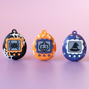 May include: Three Halloween-themed digital pet toys in black, orange, and blue. Each egg-shaped toy features a small screen displaying a Halloween icon: a crab, a jack-o'-lantern, and a witch's hat. The toys are set against a soft pink background.