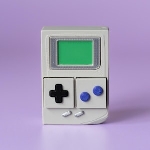 May include: A small, handheld gaming device in a light gray color, featuring a green screen and black and blue buttons. The device has a retro design, reminiscent of classic gaming consoles, and is set against a purple background.