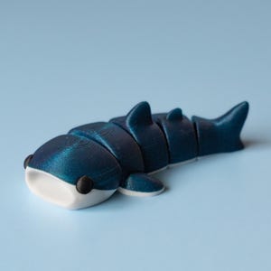 May include: A blue and white articulated whale shark toy. The toy is segmented, allowing for movement. The whale shark has a white mouth and black eyes. The background is a light blue.