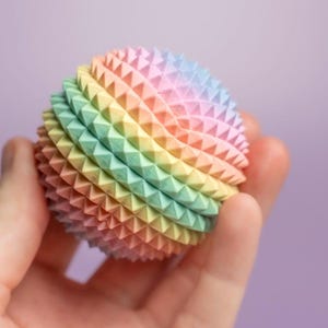 May include: A multi-coloured, spiky ball in a rainbow gradient of pastel shades, held in a hand. The ball's surface is covered in small, triangular spikes, creating a textured appearance. The background is a soft purple.