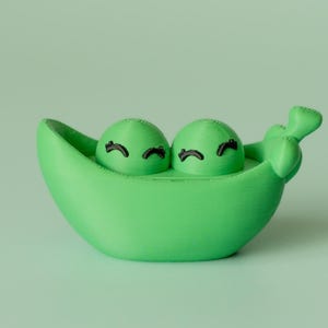 May include: A vibrant green pea pod ornament featuring two smiling peas with black eyelashes. The pod has a curved design and a stem detail. The background is a soft, light green. The item is likely 3D-printed.