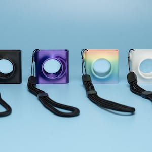 May include: Four square camera cases in black, purple, rainbow, and white, each with a circular opening and a black wrist strap. The cases are arranged on a light blue surface.