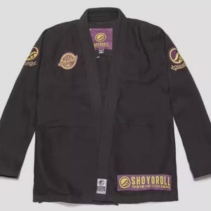 Puede incluir: Gi de Jiu-Jitsu negro con detalles dorados y morados. El gi presenta el nombre de la marca "SHOYOROLL" en letras doradas sobre un parche morado. Parches dorados con diseños están en las mangas y el pecho.