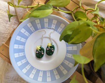 May include: Handmade green pickle earrings with yellow flower details and black facial features. The earrings are on a white plate with a blue patterned rim. A wooden tray and green plant are in the background.