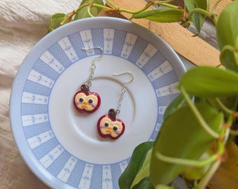 May include: Handmade apple-shaped earrings. Each earring has a red apple design with a yellow face and a silver hook. The earrings are presented on a white plate with a blue patterned rim.