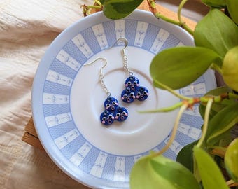 May include: Blue cluster earrings with pink details and silver hooks sit on a white plate with a blue patterned border. The earrings are made of three joined circles. Green leaves and a wooden tray are in the background.