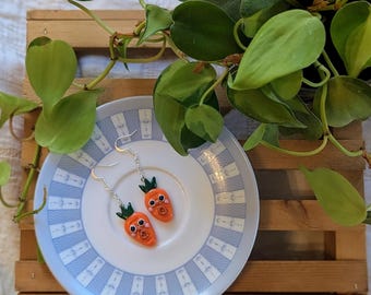 May include: Carrot-shaped earrings with silver hooks, featuring orange bodies, green tops, and small, round eyes. The earrings are displayed on a white plate with a blue patterned border, with greenery in the background.