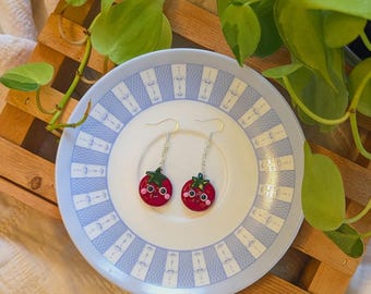 May include: Two red tomato earrings with green tops and silver hooks, each with a smiling face featuring black eyes and pink cheeks. The earrings are presented on a blue and white patterned plate, with a wooden slatted tray in the background.