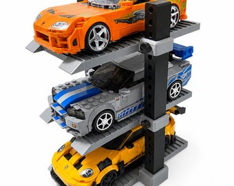 Può includere: Un espositore per parcheggio a tre livelli realizzato con mattoncini di plastica contenente tre macchinine. Il livello superiore presenta un'auto sportiva arancione, il livello centrale un'auto argentata con strisce blu e il livello inferiore un'auto sportiva gialla.