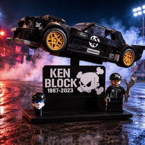 May include: A black Lego model of a Ford Hoonigan car, with gold wheels, suspended above a black base. The base features a skull logo, the text "KEN BLOCK 1967-2023", and a Lego figure holding a wrench. Smoke and stadium lights are in the background.