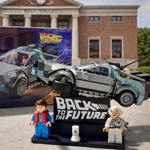 May include: A Lego Back to the Future DeLorean time machine model with two Lego figures and a sign that says "Back to the Future". The model is silver and black, with clear blue accents. The figures are Marty McFly and Doc Brown.