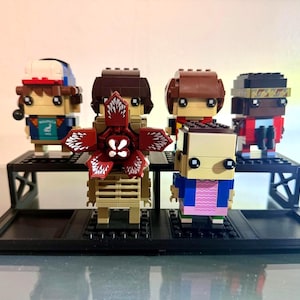 Puede incluir: Un conjunto de figuras de bloques de la serie Stranger Things, que incluye personajes como Dustin, Mike y Eleven, junto con un Demogorgon. Las figuras se exhiben en una plataforma negra escalonada, mostrando sus diseños detallados y colores vibrantes.