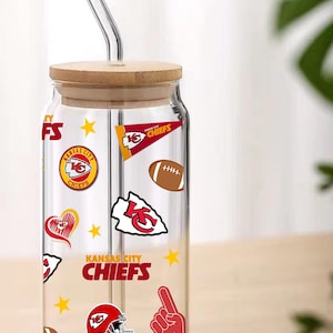May include: A clear glass tumbler with a bamboo lid and a glass straw. The tumbler is decorated with Kansas City Chiefs logos, including a red helmet, footballs, and the team name. The tumbler is designed for cold beverages.