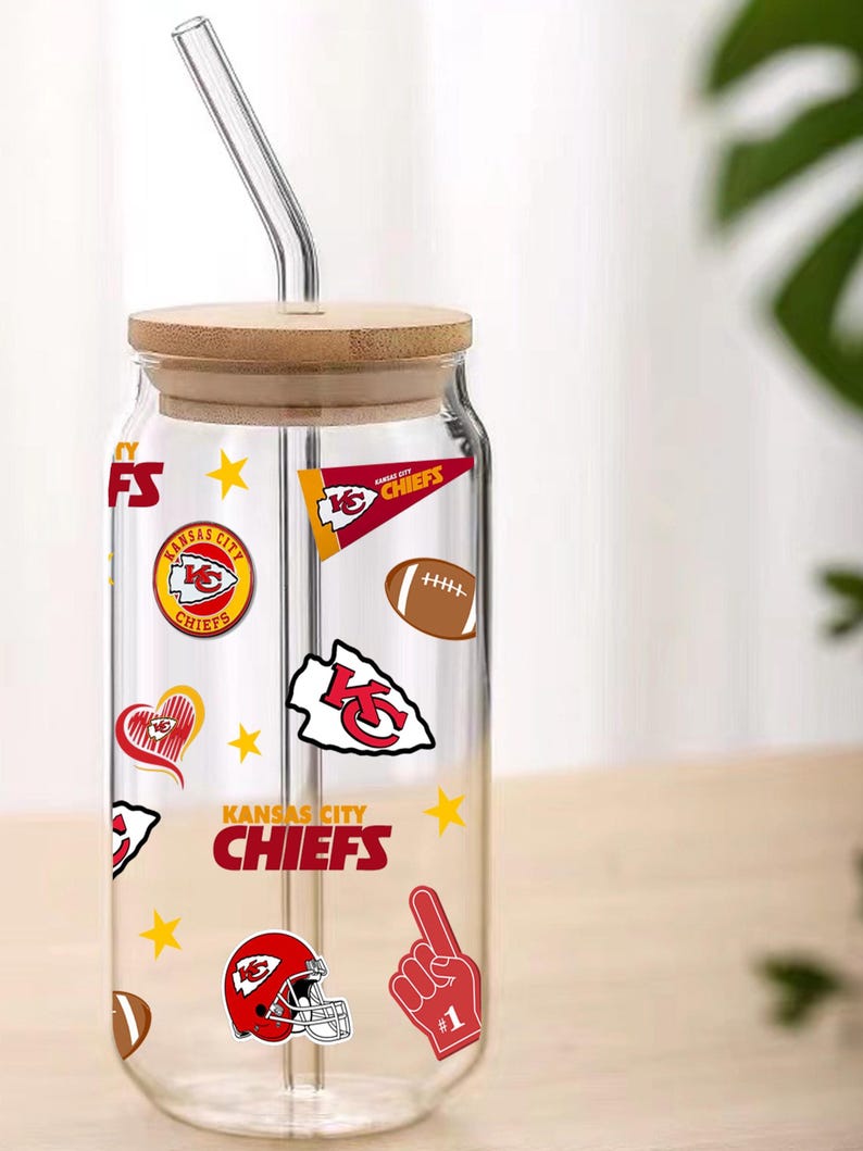 May include: A clear glass tumbler with a bamboo lid and a glass straw. The tumbler is decorated with Kansas City Chiefs logos, including a red helmet, footballs, and the team name. The tumbler is designed for cold beverages.