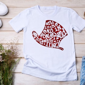 May include: White t-shirt with a red graphic of a hat with the text "and in the end, in wonderland we both went mad..."